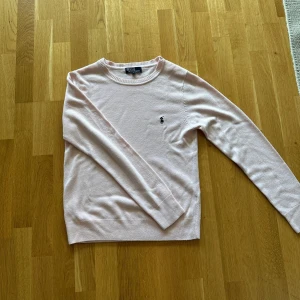 Ljusrosa stickad tröja från Polo Ralph Lauren - Supersnygg ljusrosa stickad tröja från Polo Ralph Lauren med klassisk rund halsringning och broderad logga på bröstet. Tröjan har långa ärmar och ribbade muddar vid ärmslut och nederkant. Perfekt för lager-på-lager och chill stil.