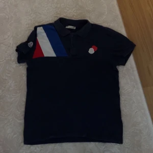 Moncler piké med färgblock och logga - Snygg mörkblå pikétröja från Moncler med coola färgblock i rött, vitt och blått på axeln. Klassisk krage och korta ärmar, samt Moncler-logga på ärmen och bröstet. Perfekt för dig som gillar sportig och stilren design.