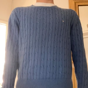 Ljusblå kabelstickad tröja från GANT - Säljer en ljusblå kabelstickad tröja från GANT med broderad logga på bröstet. Tröjan har rund halsringning och langa ärmar. Perfekt för dig som gillar klassisk och stilren design.