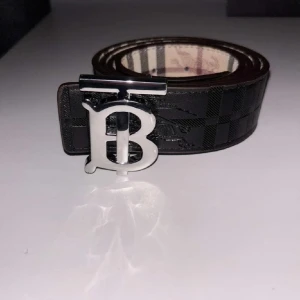 Burberry svart rutigt skärp med B-spänne - Snyggt svart skärp från Burberry med klassiskt rutigt mönster och ett stort silvrigt B-spänne framtill. Insidan har den ikoniska Burberry-rutan i beige, svart och rött. Skärpet är gjort i skinn och ger en lyxig känsla till outfiten.