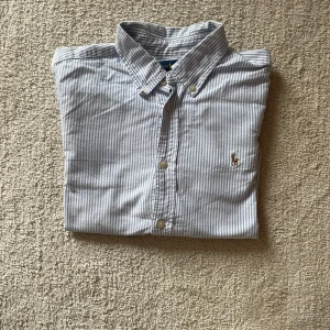 Randig skjorta från Ralph Lauren - Klassisk skjorta från Ralph Lauren i vitt och ljusblått med tunna ränder. Skjortan har button-down krage, långa ärmar och broderad logga på bröstet. Tillverkad i mjuk bomull och stängs med knappar framtill. Perfekt för en clean och stilren look.