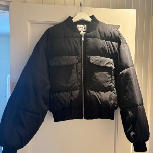 Svart pufferjacka från NA-KD, stl 36 - Säljer en svart pufferjacka från NA-KD i storlek 36. Jackan har en croppad passform, två stora fickor framtill med lock och tryckknapp, samt dragkedja hela vägen. Ribbad mudd vid krage, ärmslut och nederkant. Perfekt för kyliga dagar. använd en gång.