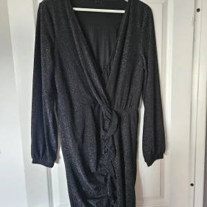 Svart glittrig omlottklänning - Svart glittrig omlottklänning med v-ringning och långa ärmar. Klänningen har en snygg rynkad detalj framtill och lätt volang längs omlottkanten. Perfekt för dig som vill sticka ut med stilren glamour.