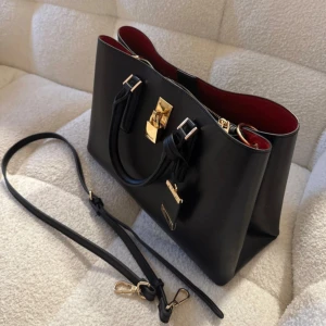 Svart handväska med gulddetaljer - Väskan har både handtag och avtagbar axelrem, samt praktiskt innerfack med dragkedja. Inga tecken på slitage då jag knappt har använt den.