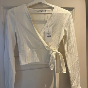 Vit omlottopp med knyt från NA-KD - Säljer en fräsch vit omlottopp från NA-KD i ribbat material. Toppen har långa ärmar, v-ringning och knyt i sidan för en snygg passform.