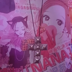 Silvrigt korshalsband med nitar - Ett halsband med ett stort korsformat hänge i silvrig metall. Hänget är designat med fyra pyramidformade nitar som ger en edgy och alternativ vibe. Hängs på ett svart snöre. Perfekt för dig som gillar statement-smycken och vill sticka ut.