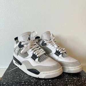 Nike Air Jordan 4 White/Black - Säljer ett par Nike Air Jordan 4 i vitt, svart och grått. Klassisk siluett med meshpaneler, snörning och detaljer i skinn och mocka. Ikonisk Jumpman-logga på plösen och hälen. Perfekta för dig som vill sticka ut med en tidlös sneaker.