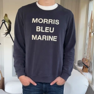 Marinblå sweatshirt från Morris - Säljer en marinblå sweatshirt från Morris med vit text 'MORRIS BLEU MARINE' på bröstet. Tröjan har rund hals, långa ärmar och ribbade muddar. Tillverkad i mjuk bomull, perfekt för en chill och stilren look.
