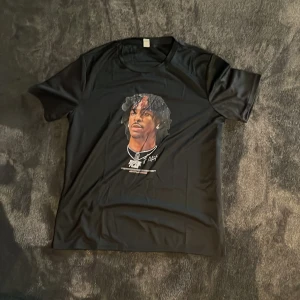 Svart t-shirt med tryck från Popular Brand - Svart t-shirt med coolt tryck av en person med halskedjor och signaturdetaljer på bröstet. Rund hals och korta ärmar, tillverkad i mjukt material som känns skönt mot huden. Perfekt för dig som gillar streetwear och grafiska motiv. Det är en träningströja!