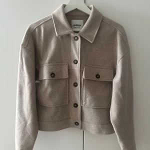 Beige jacka från Dobber - Stilren beige jacka från Dobber med klassisk krage, stora bröstfickor med knappar och raka axlar. Jackan har en rak passform, knäppning framtill och är tillverkad i ett mjukt ullmaterial. Perfekt för lager-på-lager och snygg till jeans.