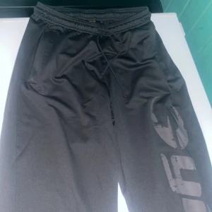 Gråa gasp byxor - Svarta mjukisbyxor med elastisk midja och snörning. Byxorna har ett stort tryck på ena benet och är tillverkade i ett mjukt material som passar perfekt för chill eller träning. Avslappnad passform och praktiska fickor på sidorna.