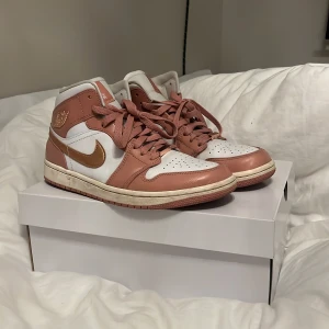 Nike Air Jordan 1 Mid rosa/vit - Säljer ett par Nike Air Jordan 1 Mid i en snygg kombo av rosa och vitt med metallic guld swoosh. Skorna har klassisk snörning, rund tå och är tillverkade i skinn. Perfekta för dig som vill sticka ut med stilrena sneakers med ikonisk design.