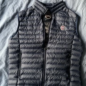 Mörkblå dunväst från Colmar - Snygg mörkblå dunväst från Colmar med quiltad design och två dragkedjefickor framtill. Västen har hög krage och Colmar-logga på bröstet. Perfekt för lager-på-lager och enkel att matcha med olika outfits. Perfekt för hösten ner du vill ha en snygg tröja men ändå hålla dig varm