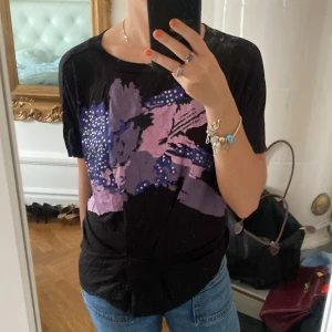 Svart t-shirt Replay med lila tryck XS - Svart t-shirt från Replay i bomull med stort abstrakt tryck i lila och blått framtill, samt små silvriga detaljer. Skön passform med korta ärmar och rund hals. Fungerar även som en kort kort klänning.