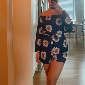 Blommig offshoulder playsuit - Säljer en superfin marinblå playsuit med stora vita blommor. Klänningen har offshoulder-modell, långa ärmar och ett knytband i midjan som ger en snygg siluett. Perfekt för sommarens alla tillfällen!