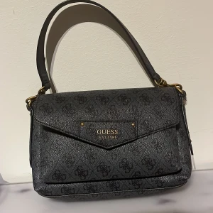 Svart axelväska från Guess med mönster - Snygg svart axelväska från Guess med diskret logomönster över hela väskan. Väskan har guldfärgade metalldetaljer, praktiskt lock med magnetknäppning och dragkedja baktill. 