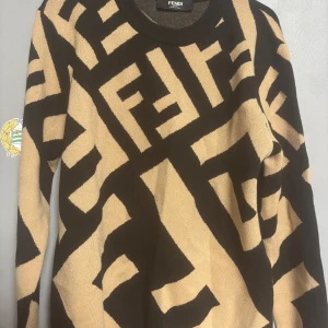 Beige och svart stickad Fendi-tröja - Säljer en beige och svart stickad tröja från Fendi med ikoniskt grafiskt FF-mönster över hela plagget. Tröjan har rund halsringning och långa ärmar. Perfekt statement piece för dig som gillar lyxiga märken och unik stil.