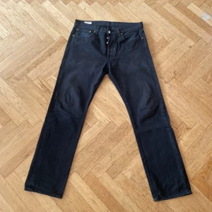 Levi's 501 svarta jeans W32 L32 - Svarta Levi's 501 jeans med klassisk rak passform och knappgylf. Jeansen har fem fickor, läderpatch bak och den ikoniska röda Levi's-taggen på bakfickan. Tillverkade i slitstarkt bomullsjeans, perfekta för en clean och tidlös look.