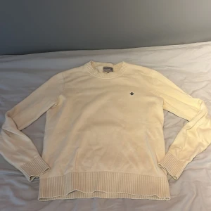 Beige stickad tröja från Morris - Snygg beige stickad tröja från Morris med rund hals och diskret svart broderad logga på bröstet. Tröjan har ribbade muddar vid ärmslut och nederkant samt långärmad passform. Perfekt för lager-på-lager och stilrena outfits.
