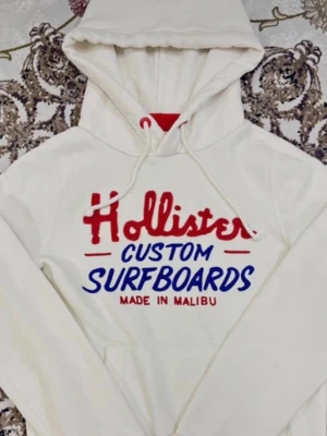 Vit Hollister hoodie med tryck - Vit hoodie från Hollister med rött och blått tryck på bröstet där det står 'Hollister Custom Surfboards Made in Malibu'. Tröjan har huva med snörning, magficka och långa ärmar. Perfekt för en chill och sportig stil. De är bara att ställa frågor!:)