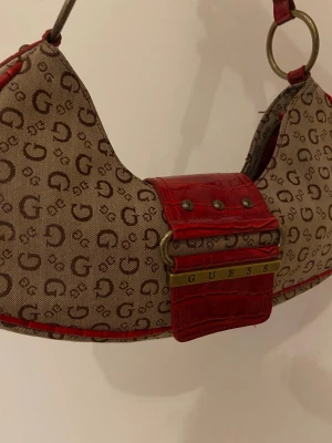 Beige och röd axelväska från Guess - Säljer en snygg axelväska från Guess med beige canvas och brunt G-mönster över hela väskan. Väskan har röda detaljer i croco-mönstrat skinn och metallringar på axelremmen. Perfekt storlek för det viktigaste och har en cool retrostil.