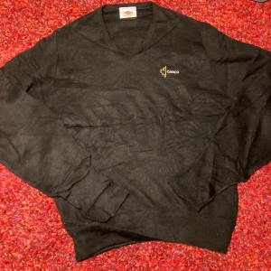 Svart v-ringad tröja från Canco - Svart v-ringad tröja från Canco med broderad logga i gult och vitt på bröstet. Tröjan har långa ärmar och ribbade muddar vid ärmslut och nederkant. Tillverkad i 100% akryl, perfekt för dig som gillar enkel och stilren design.