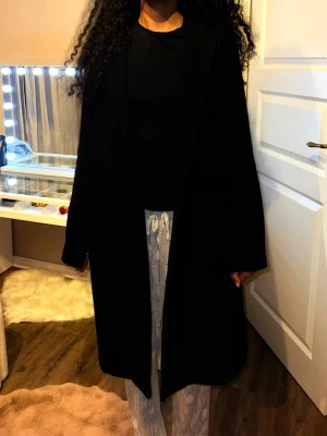 Svart lång kappa i ull - Säljer en svart, stilren och lång kappa med rak passform och lång ärm. Jackan är enkel utan synliga detaljer och har en klassisk look som funkar till många olika outfits. Materialet är mjukt och känns som ull, perfekt för kyliga dagar.