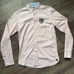 Rosa och vit randig skjorta Morris - Snygg skjorta från Morris i storlek S med tunna rosa och vita ränder. Klassisk button-down krage, broderad logga på bröstet och ljusa knappar. Tillverkad i mjuk bomull som känns skön mot huden. Perfekt för dig som gillar stilrena detaljer och diskret mönster.