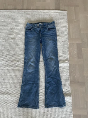 Blå bootcut jeans från Gina Tricot - Snygga blå jeans från Gina Tricot i storlek 164 med klassisk bootcut-passform. Jeansen har två framfickor, två bakfickor och stängs med dubbla silverknappar. Materialet är slitstarkt denim och färgen är en cool mellanblå ton med lätt tvättade detaljer.