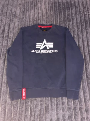 Mörkblå sweatshirt Alpha Industries - Säljer en mörkblå sweatshirt från Alpha Industries med vit logotyp tryckt på bröstet. Tröjan har långa ärmar, rund hals och en röd detalj vid nederkanten. Materialet är mjuk bomull och passformen är normal. Perfekt för dig som gillar streetwear och snygga loggor.