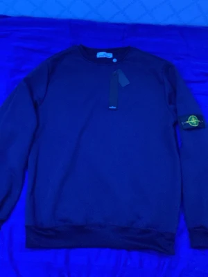 Mörkblå Stone Island sweatshirt - Säljer en mörkblå sweatshirt från Stone Island med klassisk rund hals och ribbade muddar. Tröjan har den ikoniska Stone Island-patchen på vänster ärm och är tillverkad i mjuk bomull. Perfekt för dig som gillar streetwear och snygga detaljer.