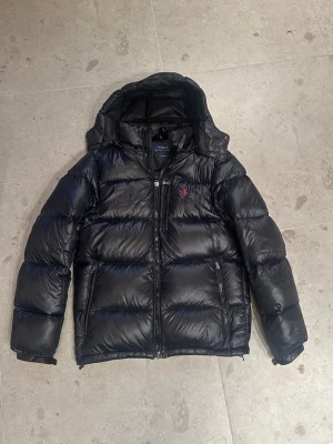 Svart jacka från Polo Ralph Lauren - Svart pufferjacka från Polo Ralph Lauren med huva och dragkedja framtill. Jackan har två sidofickor med dragkedja och ett litet rött Polo-logo broderat på bröstet. Tillverkad i återvunnen nylon och polyester, perfekt för kalla dagar.