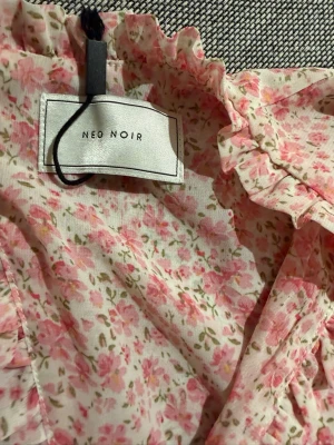 Blommig kortklänning från Neo Noir - Supersöt kortklänning från Neo Noir i ljusrosa och vit med blommigt mönster. Klänningen har volanger, v-ringning, långärmade ärmar och öppen rygg med knyt. Tillverkad i ett lätt och luftigt material, perfekt för vår och sommar. Använd endast en gång!!!