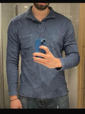Blå långärmad henley från +351 - Säljer en blå långärmad henley från +351 med mjukt bomullstyg och en chill krage. Skjortan har en bröstficka och avslappnad passform, perfekt till jeans. Enkel design utan mönster och med klassiska knappar vid halsen.