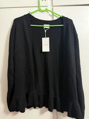 Svart kofta med volang från VILA - Svart kofta från VILA i storlek L. Koftan har långa ärmar och en öppen front utan knappar. Nedtill finns breda ribbade volanger som ger en cool och trendig touch. Materialet är stickat och känns mjukt mot huden. Perfekt att slänga på sig när det är lite kyligt ute.