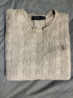 Beige stickad tröja från Polo Ralph Lauren - Säljer en beige stickad tröja från Polo Ralph Lauren med klassiskt kabelstickat mönster och rund hals. Tröjan har långa ärmar och ribbade muddar vid ärmslut och nederkant. Diskret logga broderad på bröstet. Perfekt för dig som gillar stilren och tidlös design.