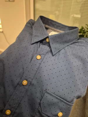 Blå prickig skjorta från Huzar Denim - Snygg blå skjorta från Huzar Denim med små svarta prickar och kontrasterande gula knappar. Regular fit med klassisk krage, bröstficka med lock och långa ärmar. Perfekt för dig som gillar stilrena detaljer och mönster.