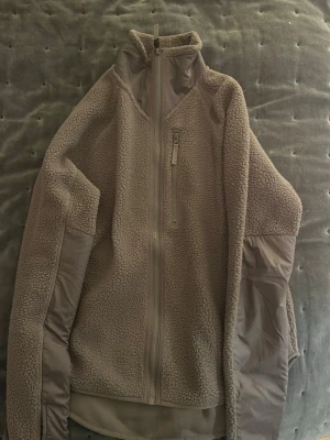 Beige teddyjacka från Move, S - Mysig beige teddyjacka från Move i storlek S. Jackan har hög krage, hel dragkedja framtill och en bröstficka med dragkedja. Ärmarna har förstärkta partier i matchande beige färg. Perfekt för kyliga dagar och har en loose passform.