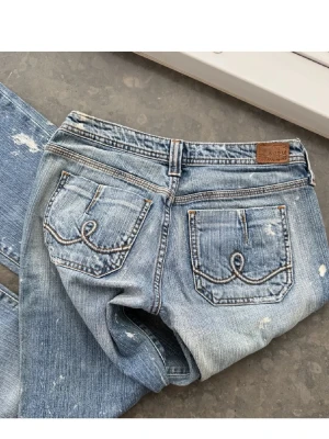 Blå bootcut jeans med slitningar - Säljer ett par ljusblå bootcut jeans med slitna detaljer och råa kanter. Jeansen har klassiska fem fickor, snygga broderier på bakfickorna och en låg midja. Perfekta för dig som gillar en avslappnad och trendig look. (lånade bilder, skriv för egna) 