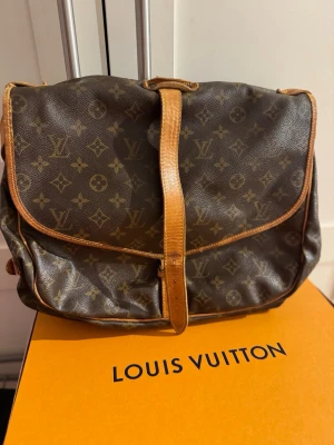 Louis Vuitton brun axelväska med monogram - Säljer en klassisk brun axelväska från Louis Vuitton med ikoniskt monogrammönster. Väskan har justerbar axelrem och snygga läderdetaljer i beige. Perfekt storlek för vardagsgrejer och har ett spänne framtill för extra stilpoäng.