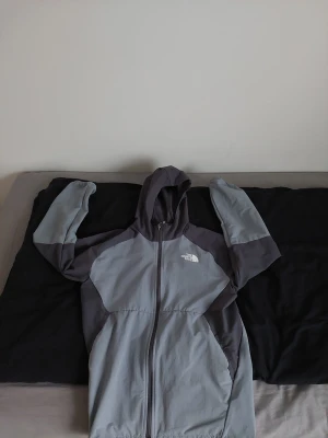 Grå vindjacka från The North Face - Säljer en snygg tvåfärgad vindjacka från The North Face i ljusgrått och mörkgrått. Jackan har huva, hel dragkedja och logga på bröstet. Perfekt för blåsiga dagar och har en sportig vibe. Materialet är polyester och jackan är långärmad. Storlek 14-16 år så runt 165-175 cm.