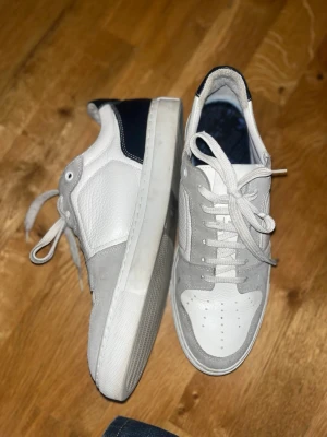 AMI Alexandre Mattiussi sneakers - Snygga sneakers från AMI Alexandre Mattiussi i vit och grå med mörkblå detaljer på hälen. Skorna har snörning, rund tå och platt sula. Ovandelen är i skinn och mocka, med AMI-logga broderad på hälen. Perfekt för dig som gillar stilrena och trendiga sneakers.