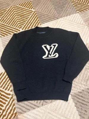 Svart stickad tröja med LV-logga - Svart stickad tröja från Louis Vuitton i storlek S. Tröjan har en vit, stor LV-logga på både fram- och baksidan. Rund halsringning och ribbade muddar vid ärmslut och nederkant. Perfekt för dig som gillar exklusiva streetwear-plagg.