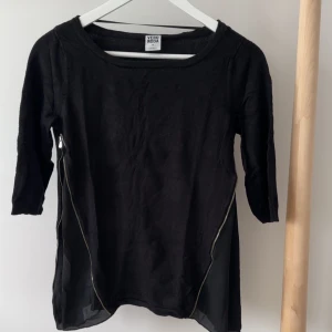 Svart tröja med dragkedjor Vero Moda - Svart tröja från Vero Moda med trekvartsärm och dragkedjor längs sidorna. Lätt material med transparenta partier nedtill som ger en snygg layering-effekt.