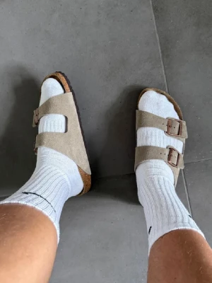 Beige Birkenstock sandaler mocka - Säljer ett par klassiska Birkenstock sandaler i beige mocka med två breda remmar och justerbara metallspännen. Sulan är ergonomisk och har en brun kant. Perfekta för chill dagar och enkel stil. Passar dig som gillar bekvämt och tidlöst.