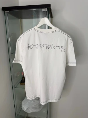Vit Acne Studios t-shirt med kontrastsömmar - Säljer en clean vit t-shirt från Acne Studios med svart broderad logga på ryggen och kontrastsömmar längs ärmar och hals. Klassisk passform och rund halsringning. Tillverkad i mjuk bomull för skön känsla. Perfekt för dig som gillar minimalistisk stil. Oversized Unisex fit 