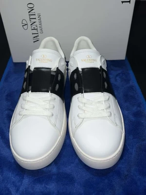 Valentino Garavani Open sneakers vit/svart - Säljer ett par Valentino Garavani Open sneakers i vitt skinn med bred svart läderdetalj över mitten och vita snören. Snygg och stilren design med rund tå och platt sula. Ikonisk logga på plösen och coola nitar på sidan. Perfekt för dig som gillar lyxiga sneakers.