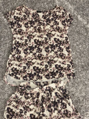 Blommig pyjamas från Emilia - Säljer en pyjamas från Emilia. Tröjan i stlk S och shortsen i stlk XS. De har ett blommigt mönster i beige, rosa och mörkblå toner. Materialet är mjukt och känns som bomull. Perfekt pyjamas som är mjuk och luftig.