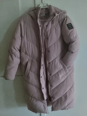 Ljusrosa lång pufferjacka från Zink - Säljer en ljusrosa pufferjacka från Zink med huva och snygga diagonala quiltade sömmar. Jackan har dragkedja och knappar framtill, två fickor och en patch på ärmen med texten 'ZINK EST. 2015'. Perfekt för kalla dagar.