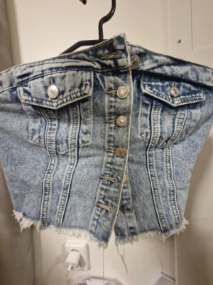 Blå denim korsett-topp med knappar - Säljer en croppad korsett-topp i ljusblå denim med snygga silverfärgade knappar framtill och två fejkfickor med lock. Toppen har råa, fransiga kanter och smockad rygg för extra stretch. Perfekt för dig som vill sticka ut med en edgy jeanslook.